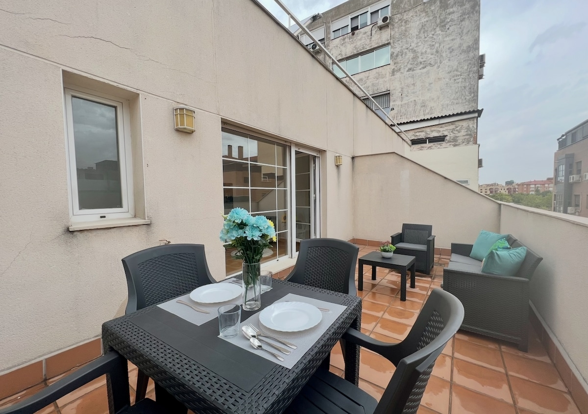 An outdoor terrace is presented with a dining table set for four. Dark wicker chairs encircle the table, which holds a vase of blue flowers. Additional seating is arranged nearby, enhancing the outdoor living space with a view of the surrounding buildings.