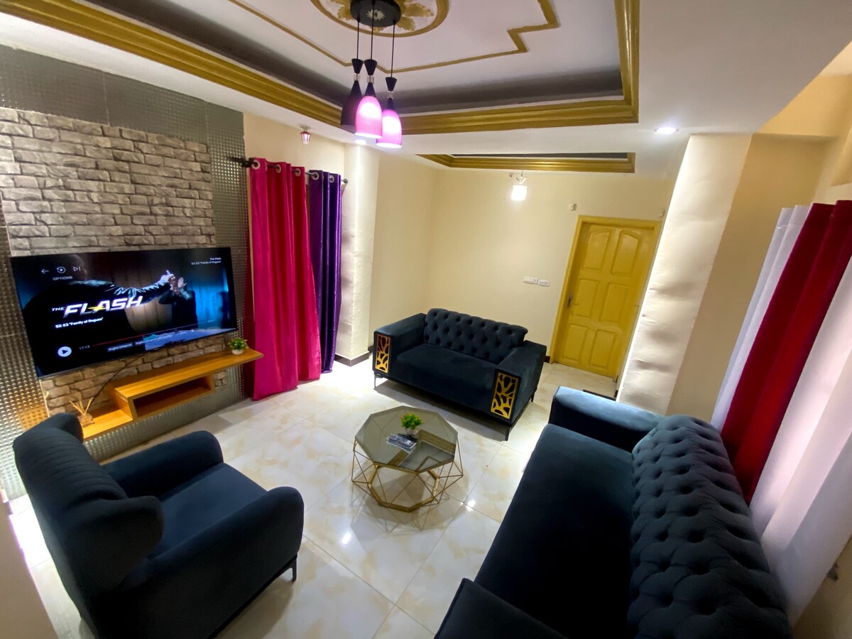 A cozy living area features dark velvet sofas arranged around a round coffee table. A large television is mounted on a textured wall, with soft lighting provided by pendant fixtures. Colorful curtains frame the windows, adding a touch of elegance to the space.