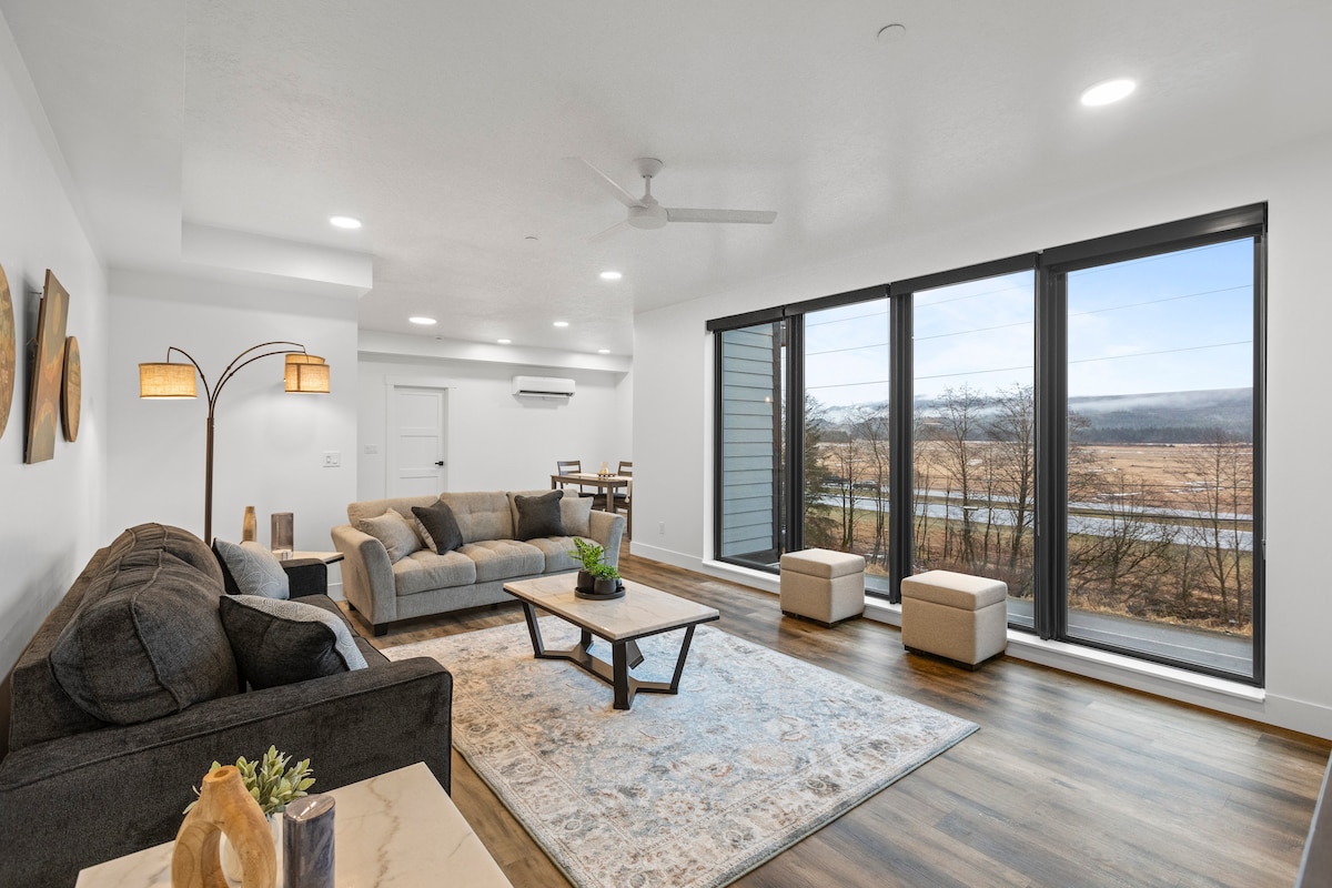 The living area features large windows that bring in natural light and offer views of the surrounding landscape. Comfortable seating is arranged around a central coffee table, while a dining area is visible in the background. A ceiling fan is mounted above for added air circulation.