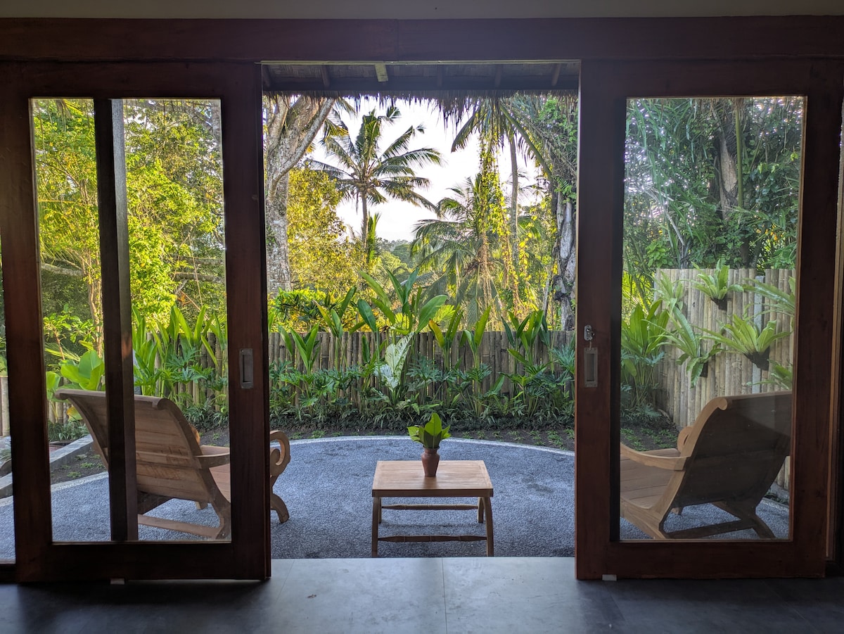Two large wooden doors are open, revealing a serene outdoor space surrounded by lush greenery. A small table sits between two wooden loungers, providing a tranquil setting to relax. The image captures the tranquility of nature, framed by the rustic architecture.