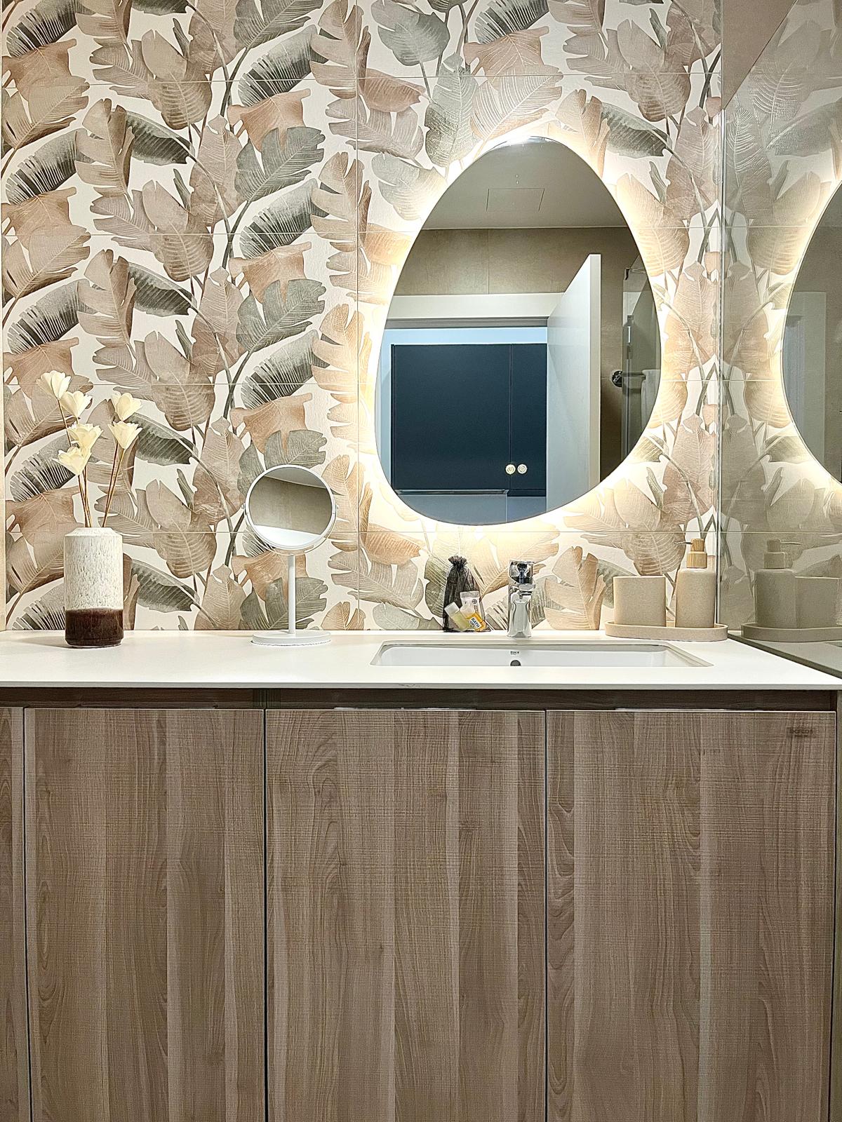A modern bathroom is highlighted by a large oval mirror with built-in lighting. The wall features a botanical print, while a wooden vanity provides storage. A small round mirror is positioned on the countertop, accentuating the clean and functional design of the space.