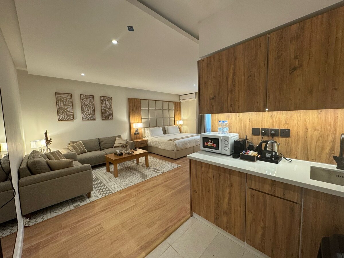 A contemporary studio apartment features a well-equipped kitchenette with modern appliances including a microwave and coffee maker. A spacious seating area with plush sofas is visible alongside a neatly made bed. Warm wood accents and soft lighting create a comfortable living space.
