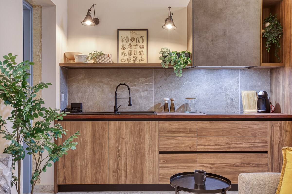 A modern kitchen area is showcased, featuring wood cabinetry and a sleek countertop. Minimalist decor includes potted plants and framed artwork on the wall. A stylish faucet and various kitchen essentials are present, complemented by warm lighting from wall-mounted fixtures.