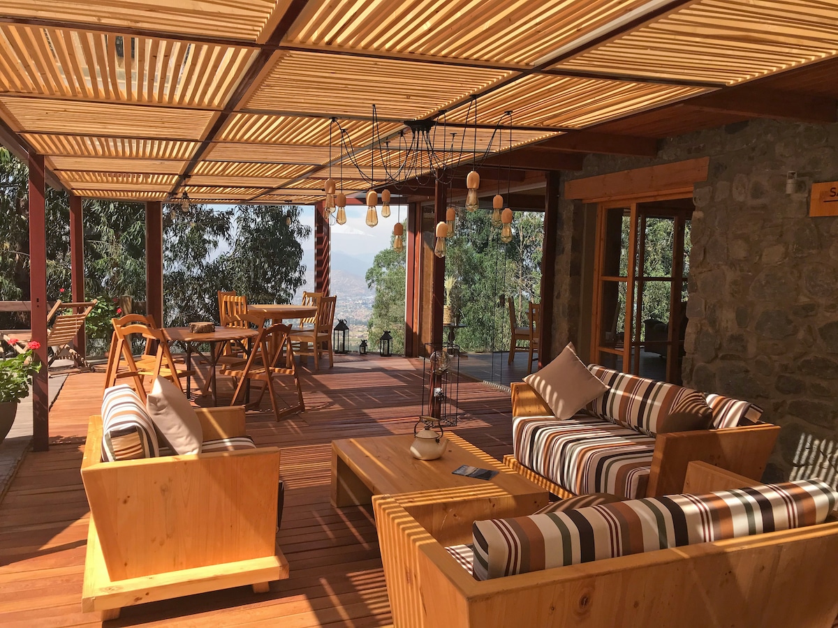 A spacious terrace area features wooden lounge furniture arranged for comfort. Natural light filters through a slatted roof, enhancing the inviting atmosphere. A dining table is set for gatherings, while surrounding greenery offers a peaceful backdrop, creating a connection with nature.