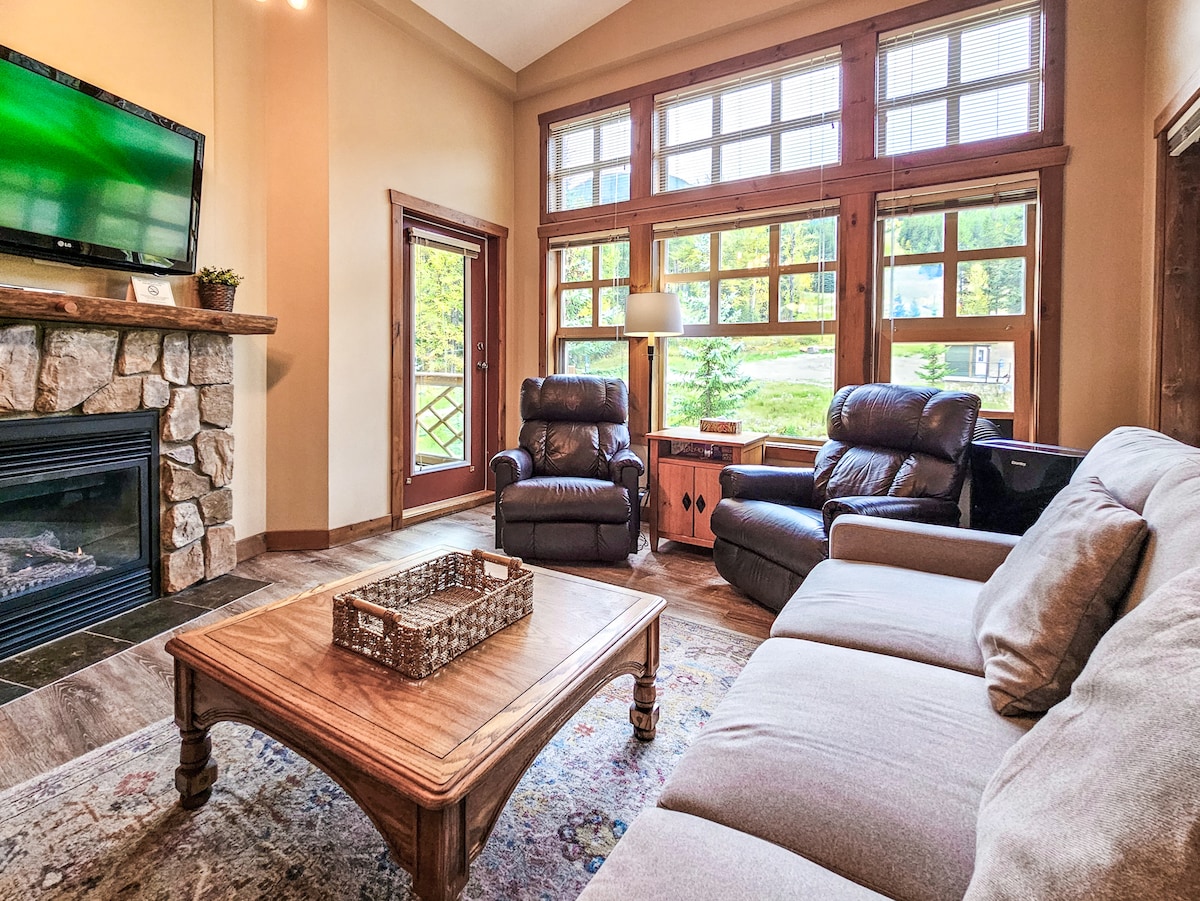 The open-plan living area features a gas fireplace framed by stone, with cozy recliner chairs and a soft sofa arranged around a wooden coffee table. Large windows allow natural light to fill the space, offering views of the outdoor landscape.