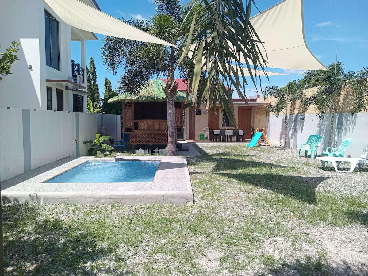 A tranquil outdoor space features a kiddie pool surrounded by sunshade sails. A kubo is situated nearby, offering shaded seating. Lush greenery and a stone pathway lead to a dining area with a table and chairs, contributing to a relaxing environment.