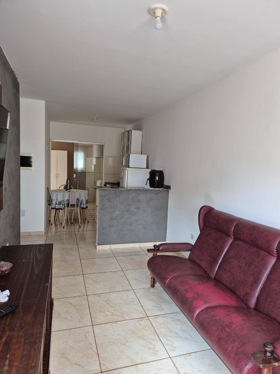A living area features a maroon sofa positioned against a light grey wall. A wooden console table is placed beside the sofa. In the background, a kitchenette is visible, showcasing white cabinets, a refrigerator, and a dining area with a small table and four chairs.