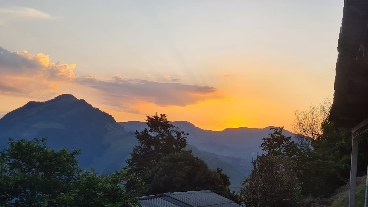 A breathtaking sunset paints the mountains in hues of orange and pink. Silhouettes of trees and rolling hills are noticeable against the colorful sky, creating a serene natural backdrop. The image captures the tranquility of the location, ideal for relaxation and enjoyment of beautiful vistas.