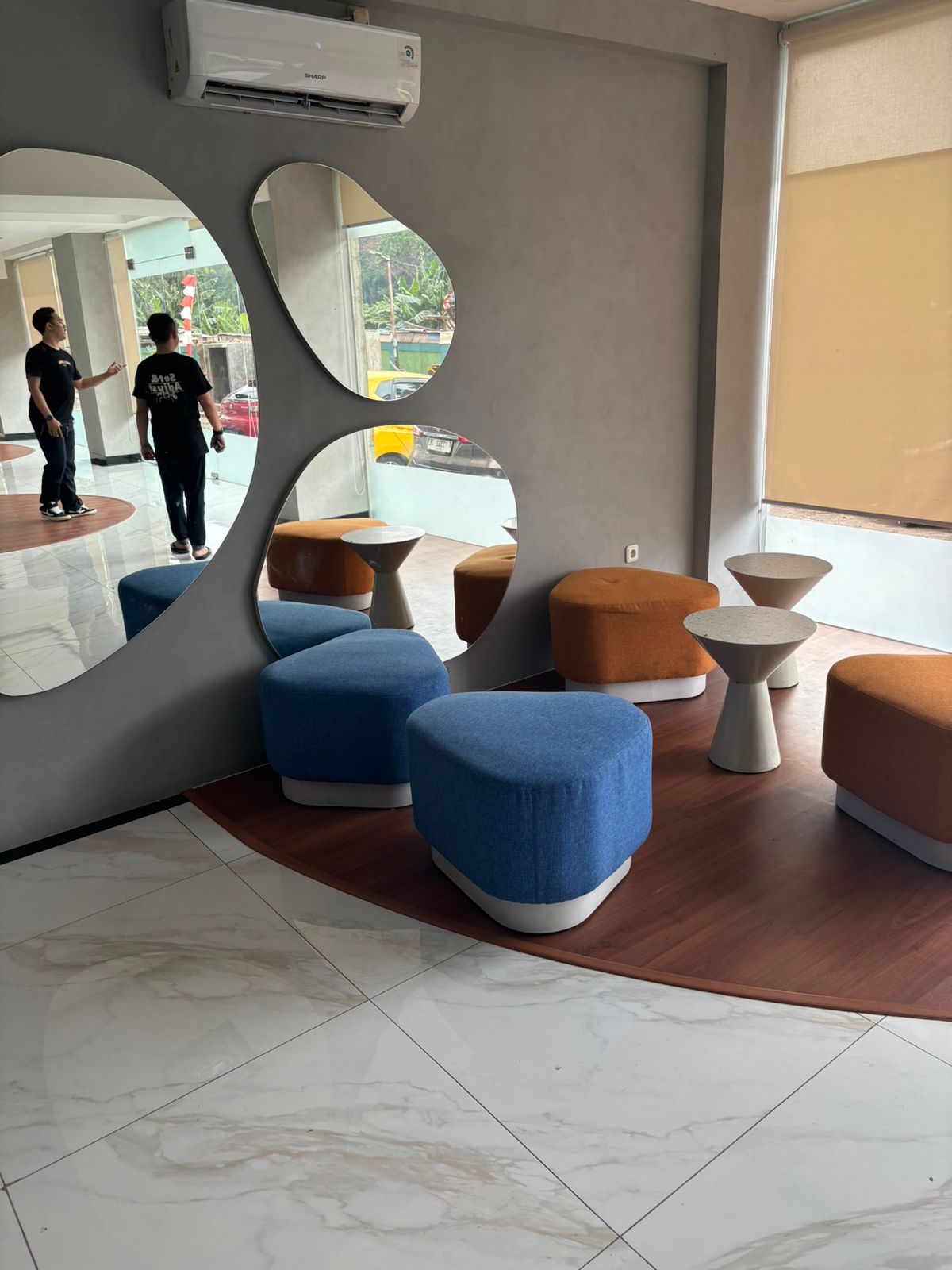 A modern seating area is highlighted by uniquely shaped upholstered chairs in blue and brown, arranged around a small white table. The space is illuminated by natural light coming through large windows, and mirrors on the wall create a sense of depth.