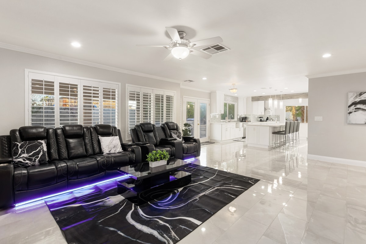 A modern living space features sleek black reclining sofas arranged around a glossy black rug. Light reflects off polished tile floors, while large windows offer views of the outdoors. The open layout extends to a bright kitchen, creating a seamless transition between areas.