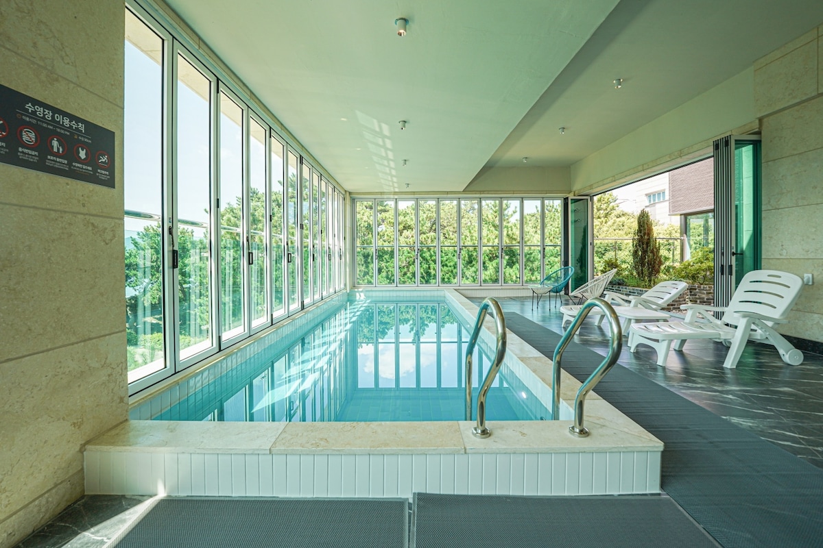 A private indoor swimming pool is highlighted, featuring large glass windows offering views of the surrounding greenery. Sun loungers are placed along the poolside, and safety signage is visible on the wall. The serene environment is complemented by natural light filtering in.
