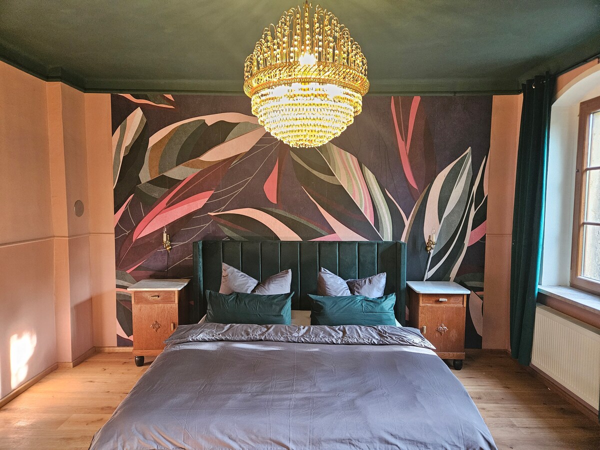 A spacious bedroom features a large chandelier with sparkling crystals. A deep green headboard frames the bed, dressed in gray linens. Colorful leaf-patterned wallpaper adds a distinctive touch, while two bedside tables stand on either side, enhancing the decorative elements of the room.