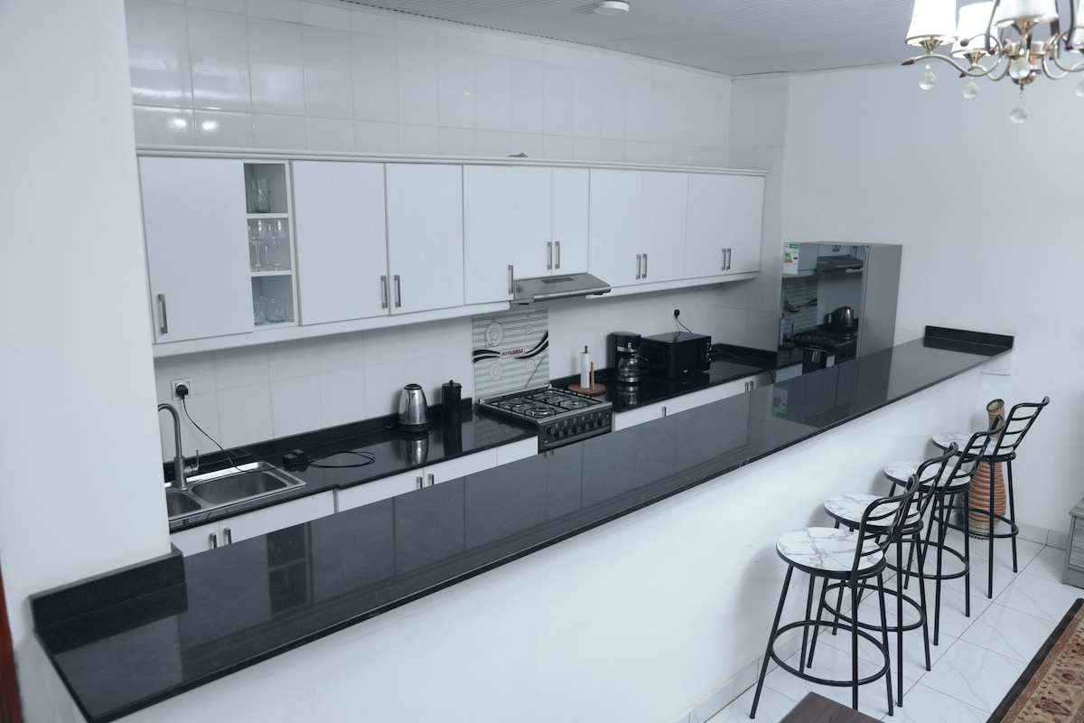 A modern kitchen is showcased with sleek white cabinetry and a black countertop. Stainless steel appliances, including a kettle and a stovetop, are visible. A bar area with three high-backed stools provides an inviting space for casual dining. Large cabinets and open shelving add functionality.