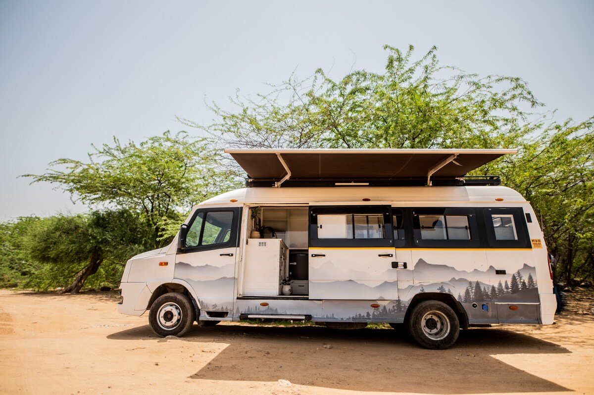 The camper van is parked on a sandy terrain, featuring a painted exterior depicting a natural landscape. A retractable awning extends from the roof, providing shade. Surrounding greenery adds a natural feel to the setting, enhancing the overall outdoor experience.