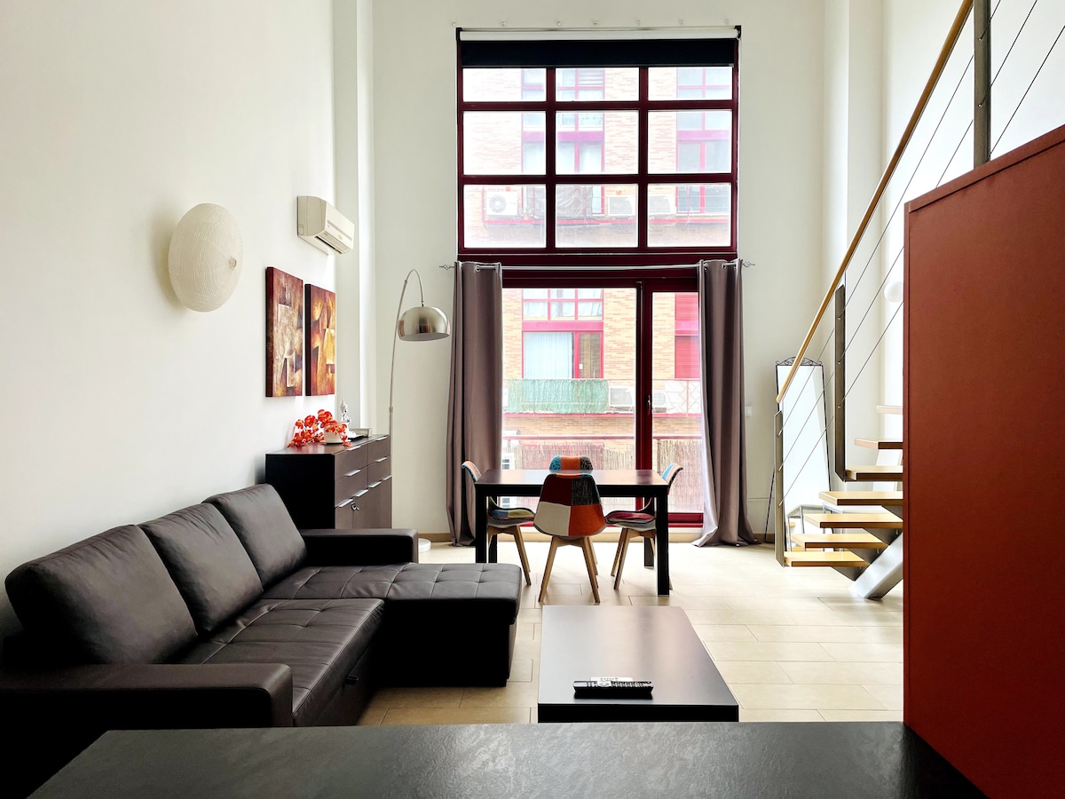 A spacious living area is presented with large windows allowing natural light to fill the space. A comfortable dark sofa is placed alongside a wooden dining table surrounded by colorful chairs. The staircase leading to the upper level adds a modern touch.