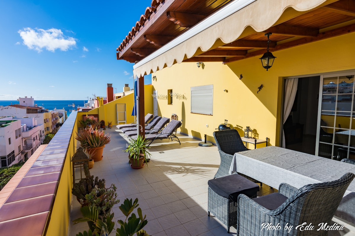 A sunlit terrace is showcased, featuring comfortable lounge chairs and a dining table. The space is surrounded by potted plants, with views of the ocean visible in the background. A warm yellow wall complements the outdoor furniture, enhancing the relaxed atmosphere.