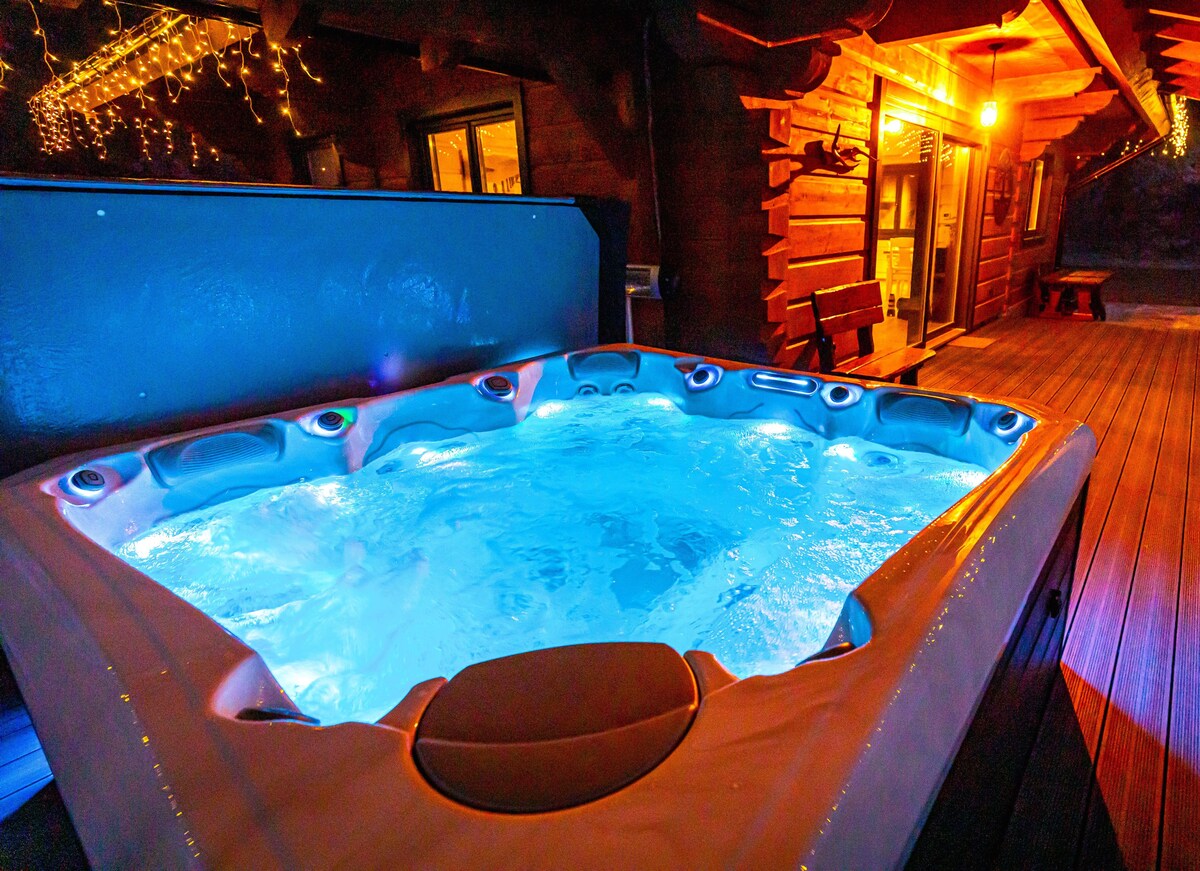 A spacious hot tub featuring soft blue lighting is situated on a wooden deck. String lights are visible above, adding a warm glow to the surroundings. The rustic wooden structure of the house can be seen in the background, enhancing the inviting outdoor space.