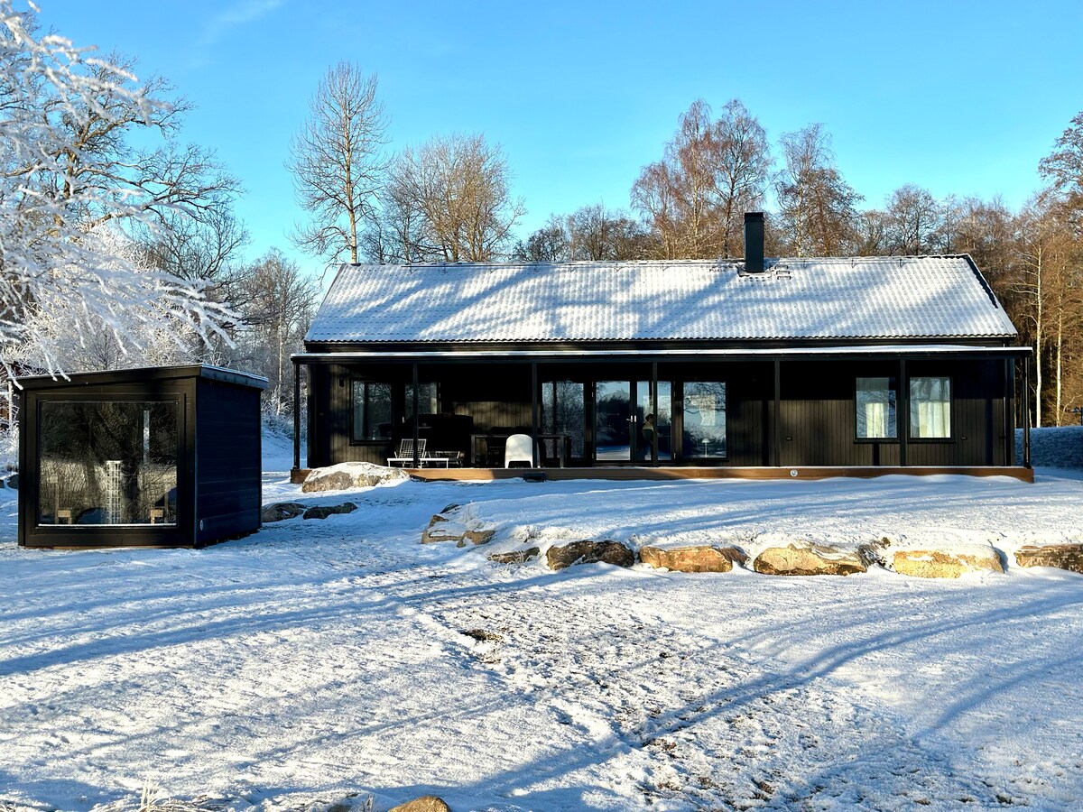 A modern black house is set against a winter landscape, surrounded by snow-covered ground and trees. Large windows allow natural light to fill the interior, while a small sauna structure is visible nearby. The serene environment offers a tranquil atmosphere.