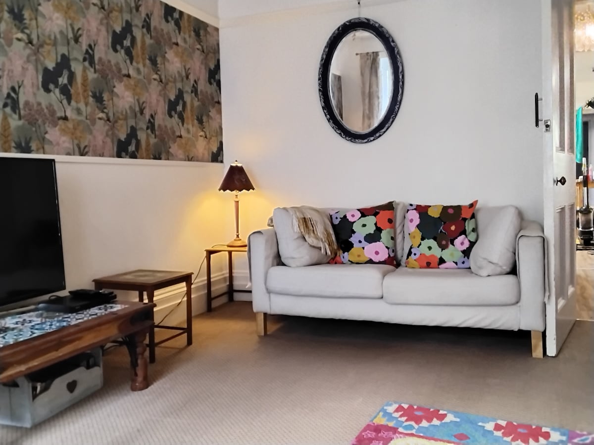 A comfortable living area features a light-coloured sofa adorned with vibrant floral cushions. A decorative mirror hangs above a small lamp and coffee table. The walls display subtle floral wallpaper, adding a touch of design. A television is positioned in view, contributing to a relaxed space.
