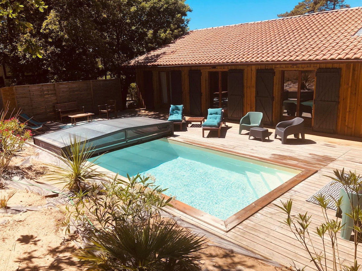The image displays a wooden terrace surrounding a private swimming pool, equipped with a safety cover. Comfortable lounge chairs and small tables are arranged nearby, offering a relaxed outdoor space. Lush greenery frames the area, creating a serene environment under clear blue skies.