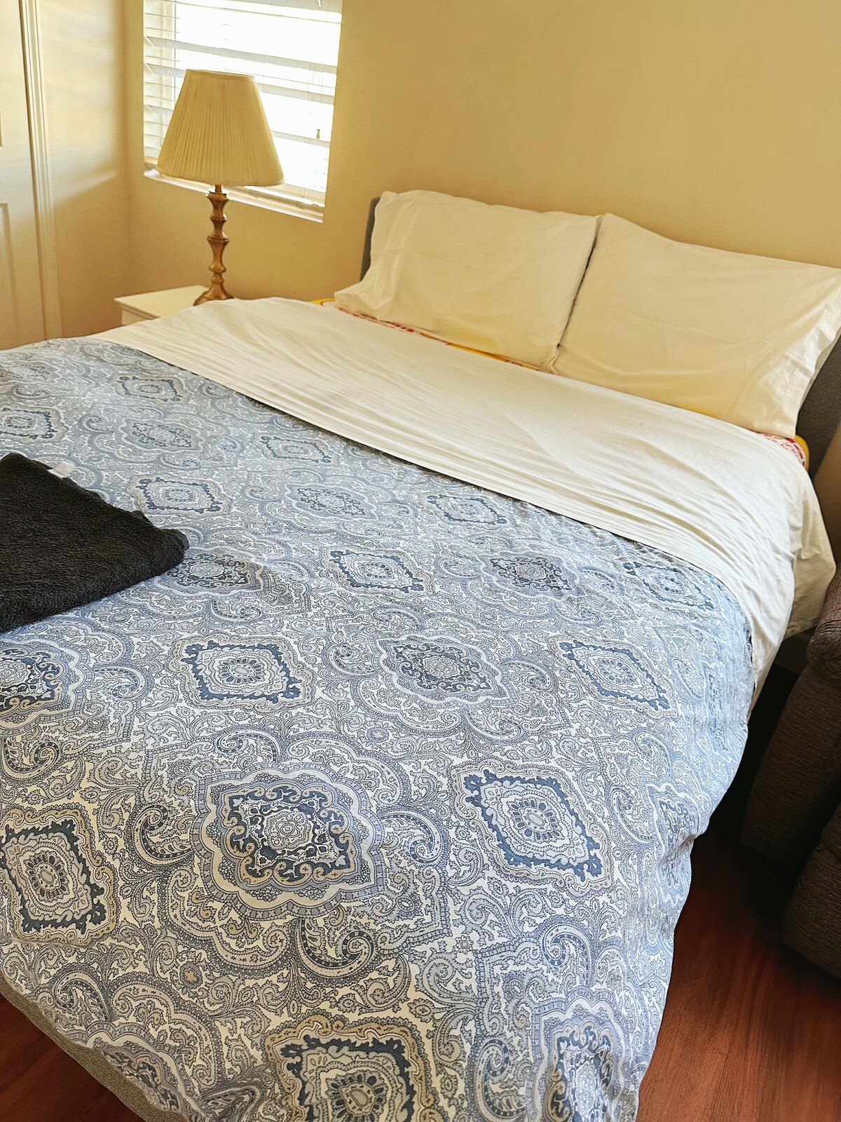 A spacious bed is dressed in a patterned blue and white cover, with a plain white sheet laid over it. A dark towel is positioned at the foot of the bed. Natural light enters through a nearby window, illuminating the room's warm tones.