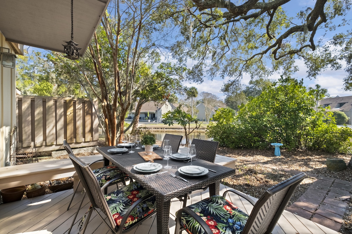 An outdoor dining area is set with a black wicker table surrounded by six chairs, decorated with tropical-patterned cushions. Lush greenery and trees form a natural backdrop, creating a serene environment. Soft sunlight filters through the leaves, enhancing the inviting atmosphere.