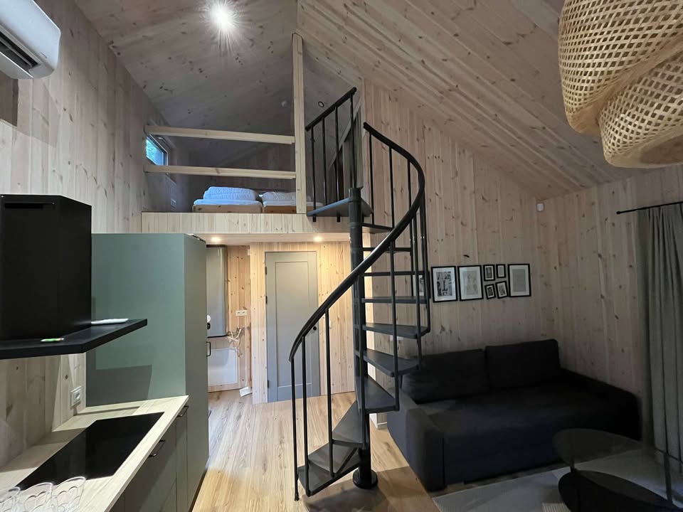 An interior view of a cozy cabin showcases a spiral staircase leading to a sleeping loft. Light wood paneling fills the walls, and a dark sectional sofa is positioned in the living area. A compact kitchen area is visible, featuring muted green cabinetry.