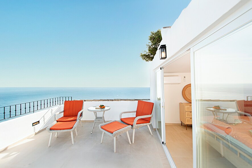 A private terrace features sunlight-washed tiles and comfortable lounge furniture. Bright orange chairs surround a small table, providing a space for relaxation. The expansive view of the sea and clear sky adds a serene backdrop, enhancing the outdoor experience.