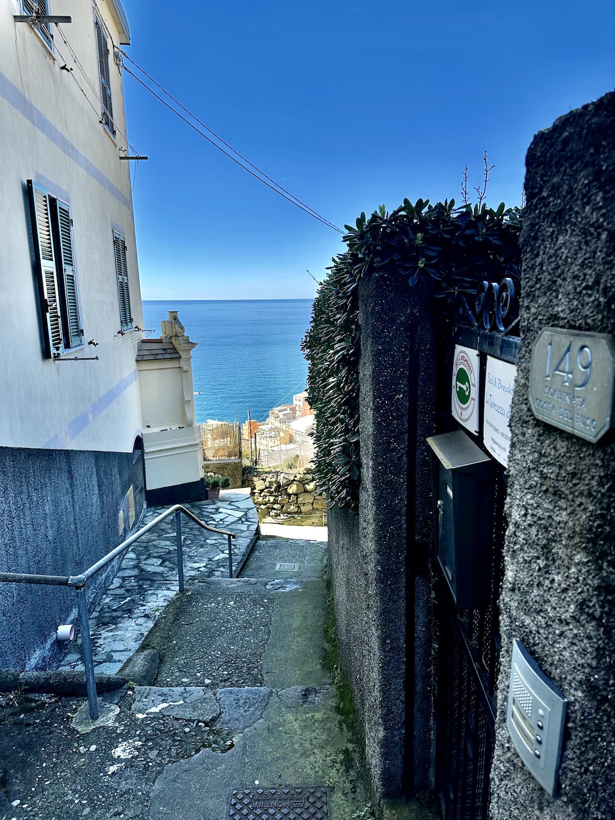 A pathway leading down from a building towards the sea is framed by stone walls and greenery. Clear blue skies and the ocean are visible at the end of the path, creating a serene coastal setting. The entrance number is displayed on the wall.