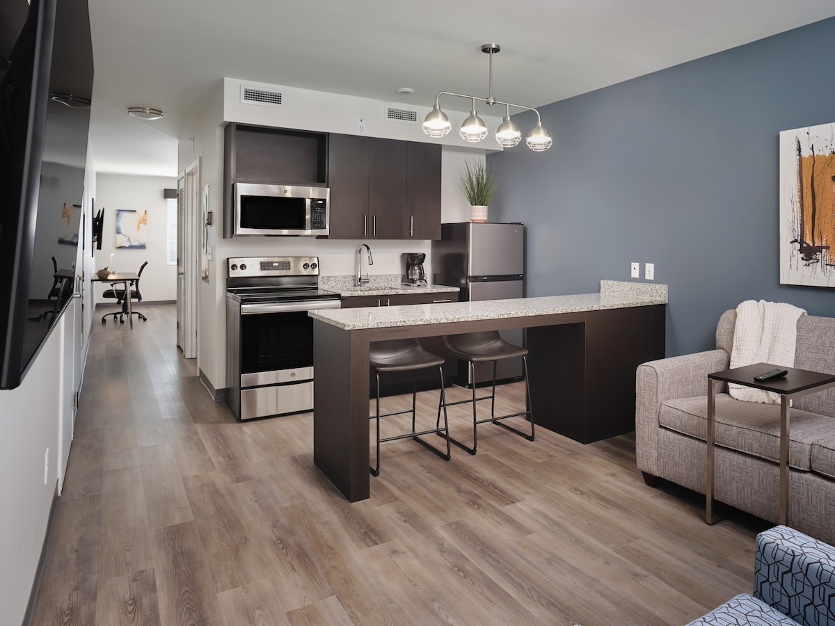 The image showcases a modern kitchen and living area, featuring stainless steel appliances, granite countertops, and a bar-height table with two stools. A comfortable gray sofa is visible, and a desk area can be seen in the background, providing a functional work space.