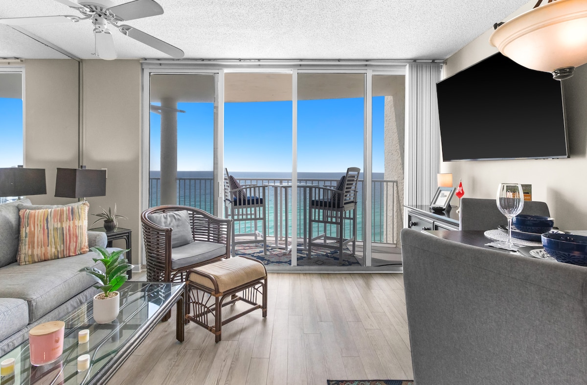 A spacious living area is highlighted by large sliding glass doors that open to a balcony overlooking the coastline. Comfortable seating is arranged around a glass coffee table, while a dining area is set with dishware. A flat-screen TV is mounted on the wall.