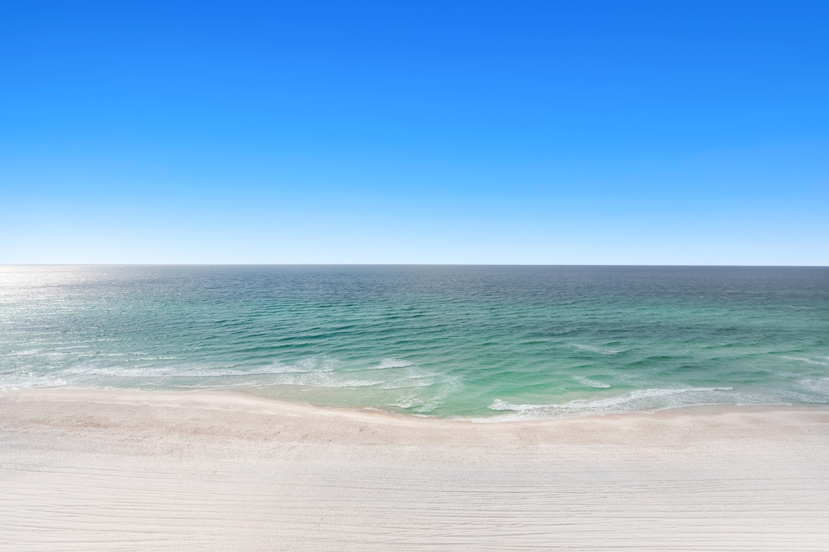 A panoramic view of the coastline is presented, showcasing azure skies meeting the gentle waves of the emerald-green ocean. The sandy beach stretches along the foreground, with soft ripples visible in the water reflecting the light of the sun.