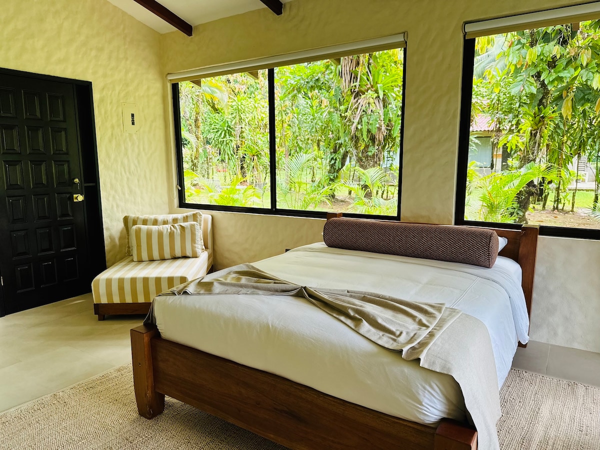 A well-appointed bedroom features a spacious bed with a light-colored sheet and a decorative bolster. A cozy chair with stripes sits beside the bed, while large windows allow natural light to illuminate the room and offer views of lush greenery beyond.