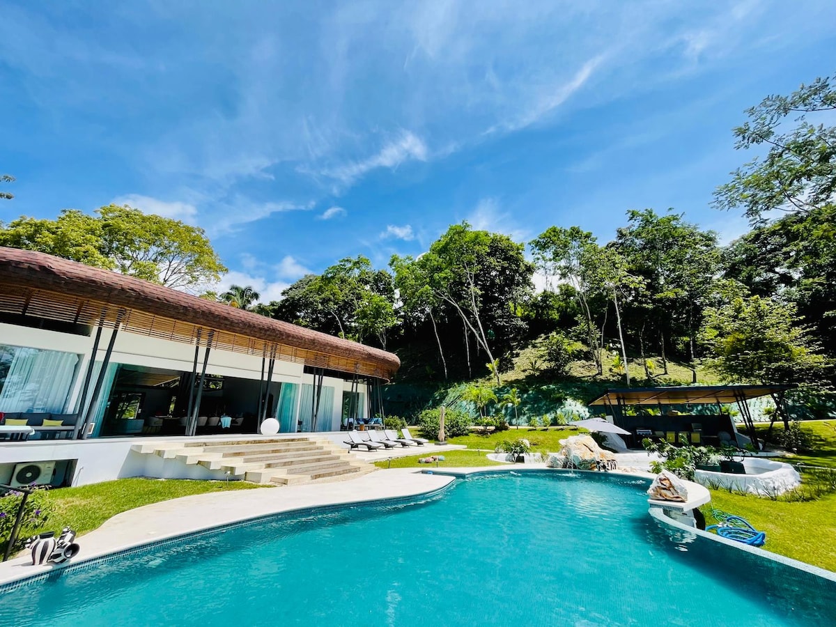 A modern home is showcased beside a large swimming pool, surrounded by lush greenery. The building features large glass windows and a thatched roof, providing an inviting connection to the outdoor environment. Lounge chairs are positioned around the pool, enhancing relaxation options.