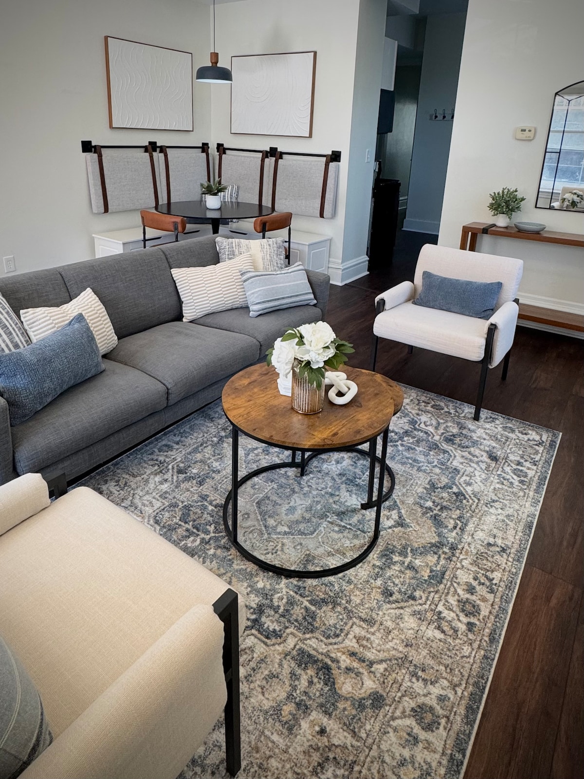 A cozy living area is arranged with a gray sofa adorned with striped pillows and accompanied by two light-colored chairs. A round wooden coffee table rests on a patterned rug, featuring a vase of flowers and decorative accents. A dining space is visible in the background.