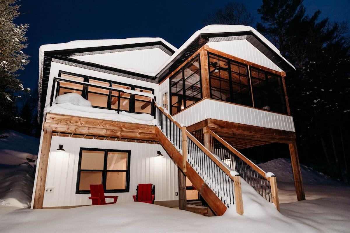 The cottage is showcased at night, with fresh snow covering the ground and surrounding elements. The exterior features a blend of white and wooden materials, along with a large staircase leading to an upper deck. Red chairs sit invitingly on the porch.