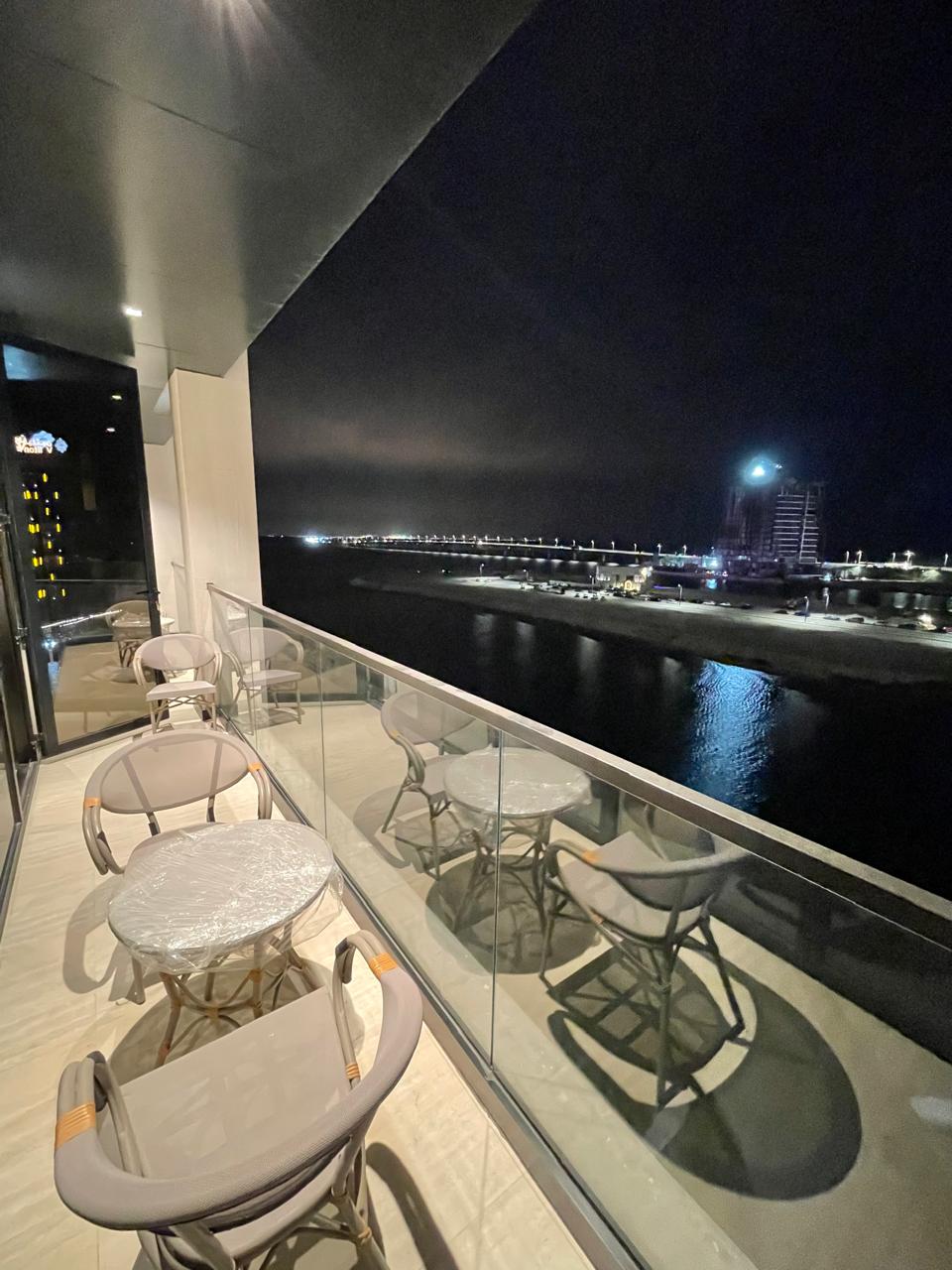 A balcony view is presented, featuring a glass railing that allows for unobstructed sightlines of the water and nearby city lights. Outdoor seating consists of four round tables accompanied by four chairs, all positioned to enjoy the serene nighttime ambiance.