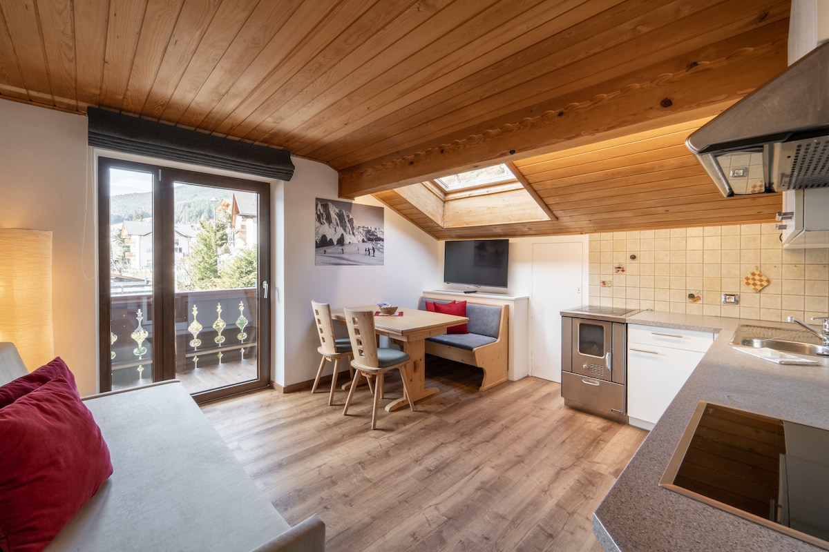 A warm living area features wooden ceilings and flooring, with a comfortable sofa positioned near a window. A dining table with chairs is set against a wall, and a kitchenette includes modern appliances. Natural light fills the space, creating a cozy ambiance.