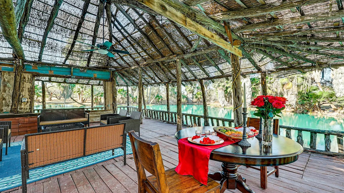 An open air pavilion is situated along a serene lake. Natural wood beams support the thatched roof, and comfortable seating areas surround a round table set with flowers and snacks. The tranquil blue water reflects the surrounding trees, creating a peaceful setting.