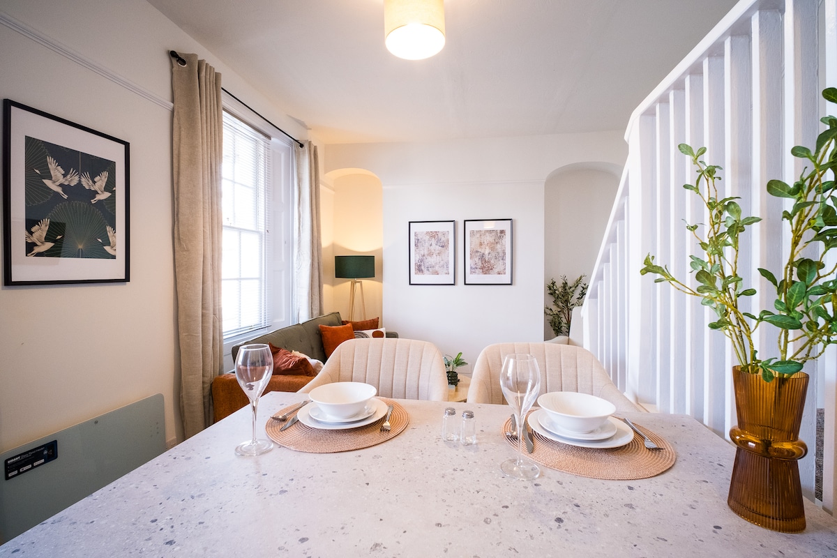 A dining area features a spacious table set for two, complete with plates and glasses. Decorative artwork is displayed on the walls, and a cozy living space is visible in the background, highlighted by natural light flowing through a large window.