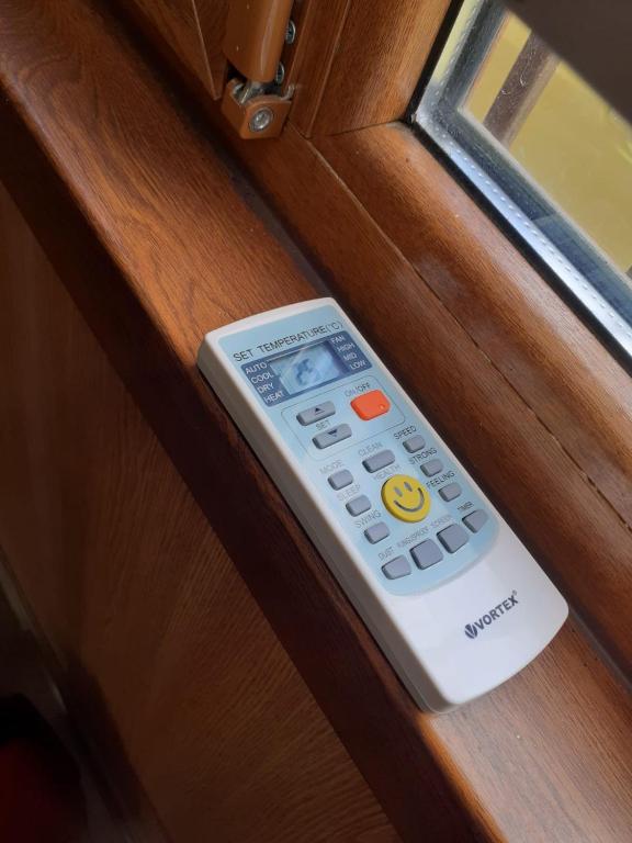 A remote control is displayed on a wooden windowsill. The device features a variety of buttons for temperature settings and functions, with a cheerful smiley face icon visible. Natural light streams through the adjacent window, highlighting the wooden texture.