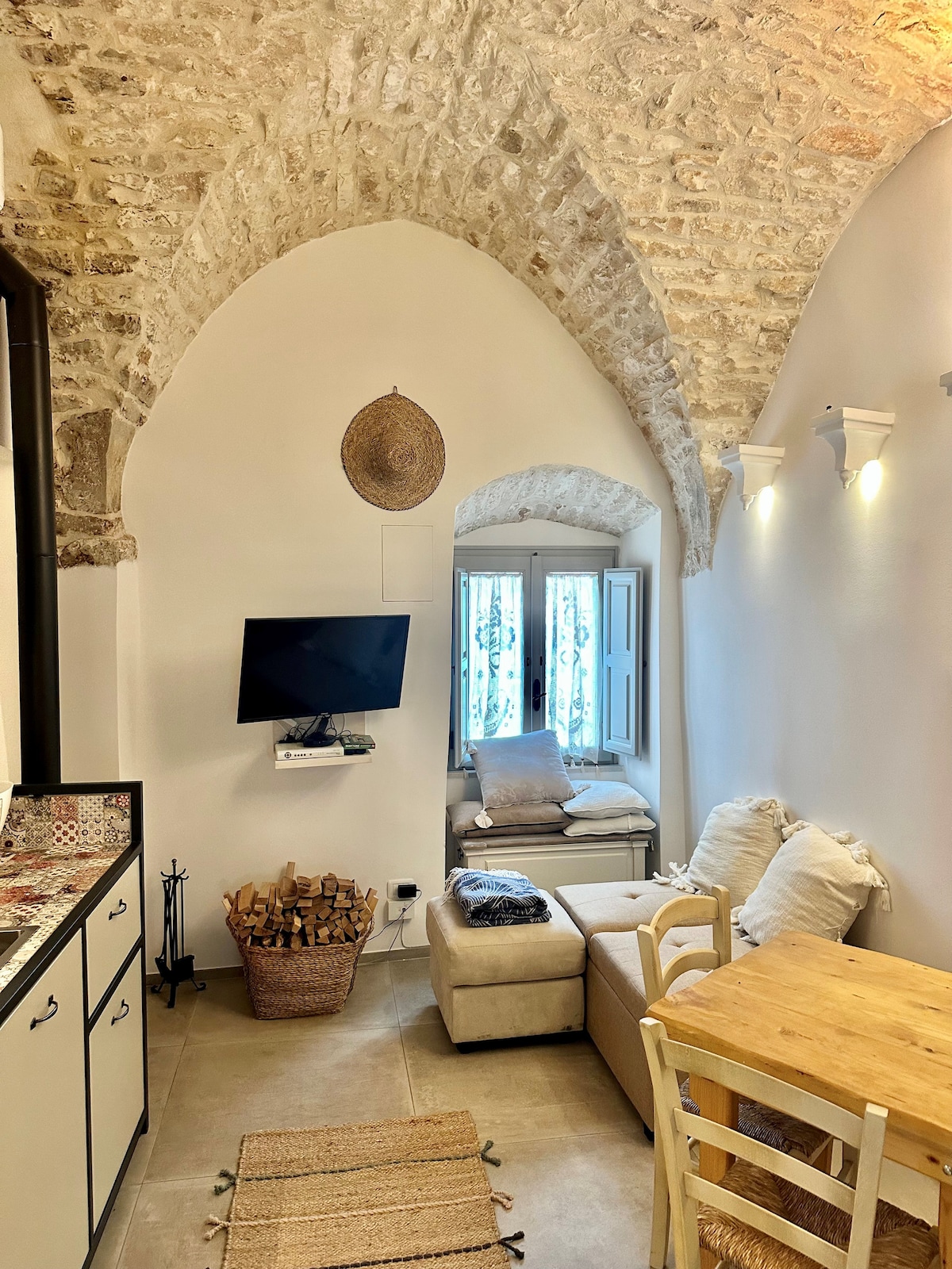 The image depicts a cozy living area with arched stone walls. A small wooden table is paired with a chair, while a comfortable couch is adorned with cushions. Natural light enters through a window with decorative shutters, enhancing the inviting ambiance.