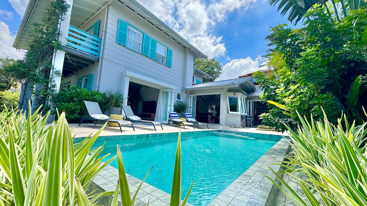 The exterior view of The Toucan House is captured, featuring a heated pool surrounded by lush greenery and tropical plants. Comfortable lounge chairs are positioned near the water, while the architecture showcases light-colored walls and blue shutters, presenting a welcoming outdoor space.