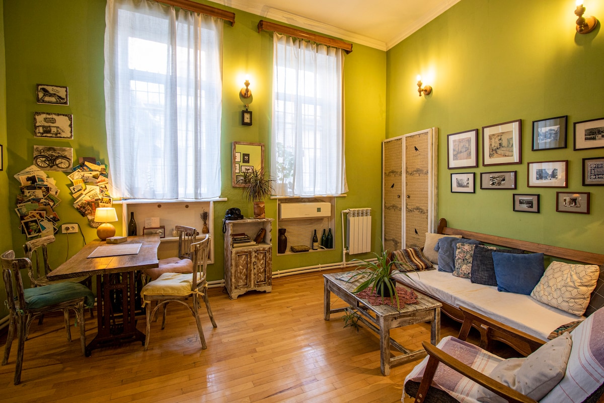 A cozy living area is enhanced by vibrant green walls and ample natural light through large windows. A wooden dining table is paired with mismatched chairs, while an upholstered sofa with colorful pillows provides comfortable seating. Vintage photographs adorn the walls, adding character to the space.