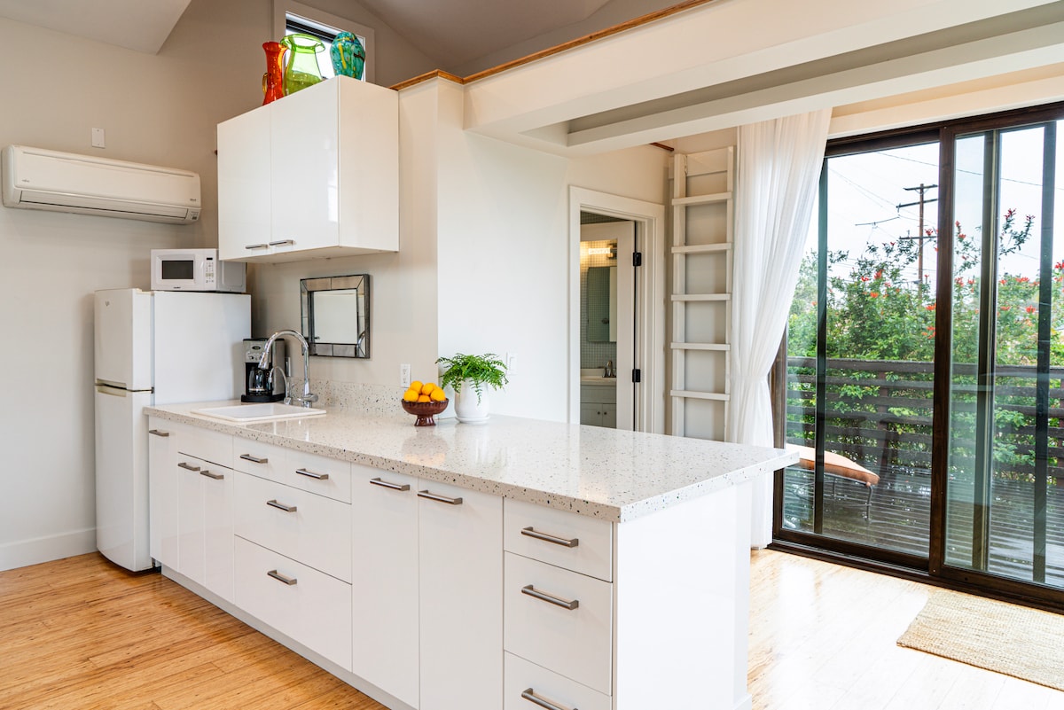 A well-equipped kitchenette features modern white cabinetry and a spacious countertop made of light-colored stone. Essential appliances including a refrigerator and microwave are visible. A window allows natural light to fill the area, while a small table with decorative fruit adds a touch of warmth.