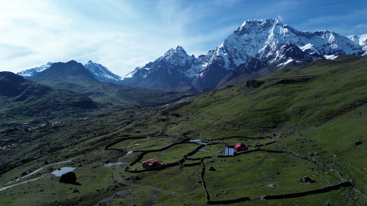 An aerial view captures the expansive landscape of lush green fields bordered by stone walls. The Mountaineers House is visible with its red roof, nestled against a backdrop of towering snow-capped mountains under a bright blue sky, framed by the rugged terrain.