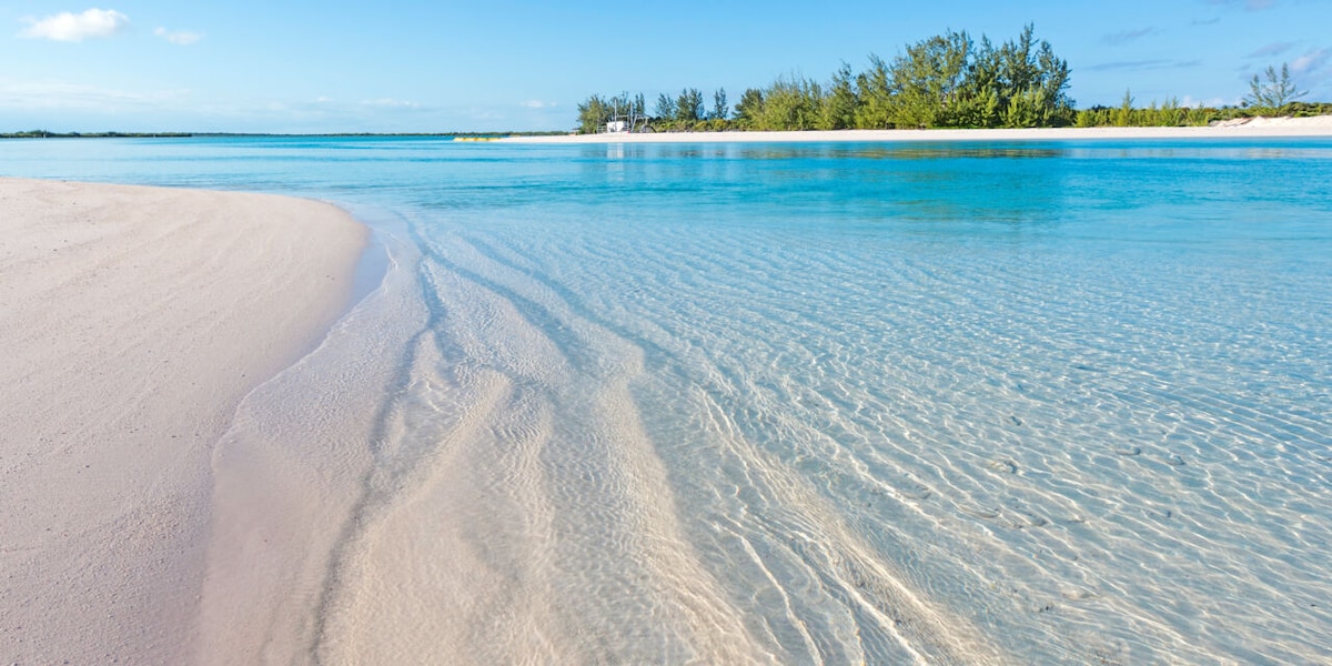A serene waterfront scene is highlighted, featuring soft, white sandy shores gently meeting clear, turquoise waters. Calm ripples create a subtle texture on the water's surface, while lush greenery marks the shoreline in the background, contributing to a tranquil coastal atmosphere.