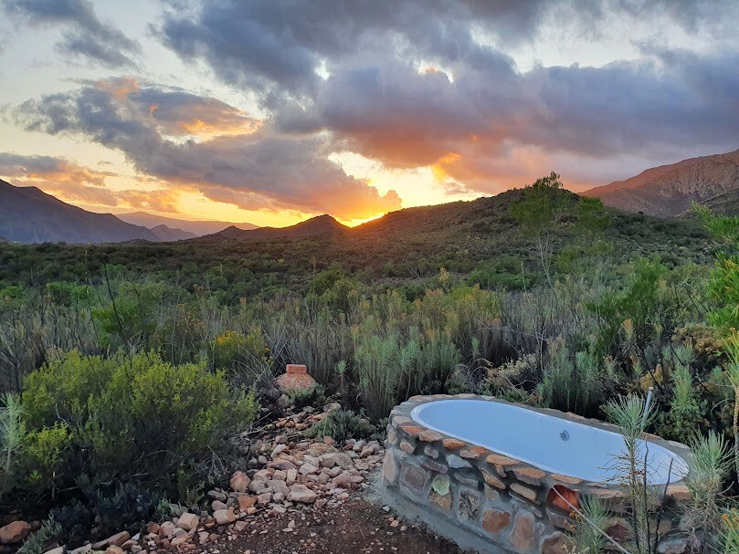 An outdoor bathtub is framed by natural stone, positioned against a backdrop of rolling hills. The setting sun casts warm hues across the sky, creating a serene atmosphere. Lush greenery surrounds the area, emphasizing the peaceful connection to the landscape.