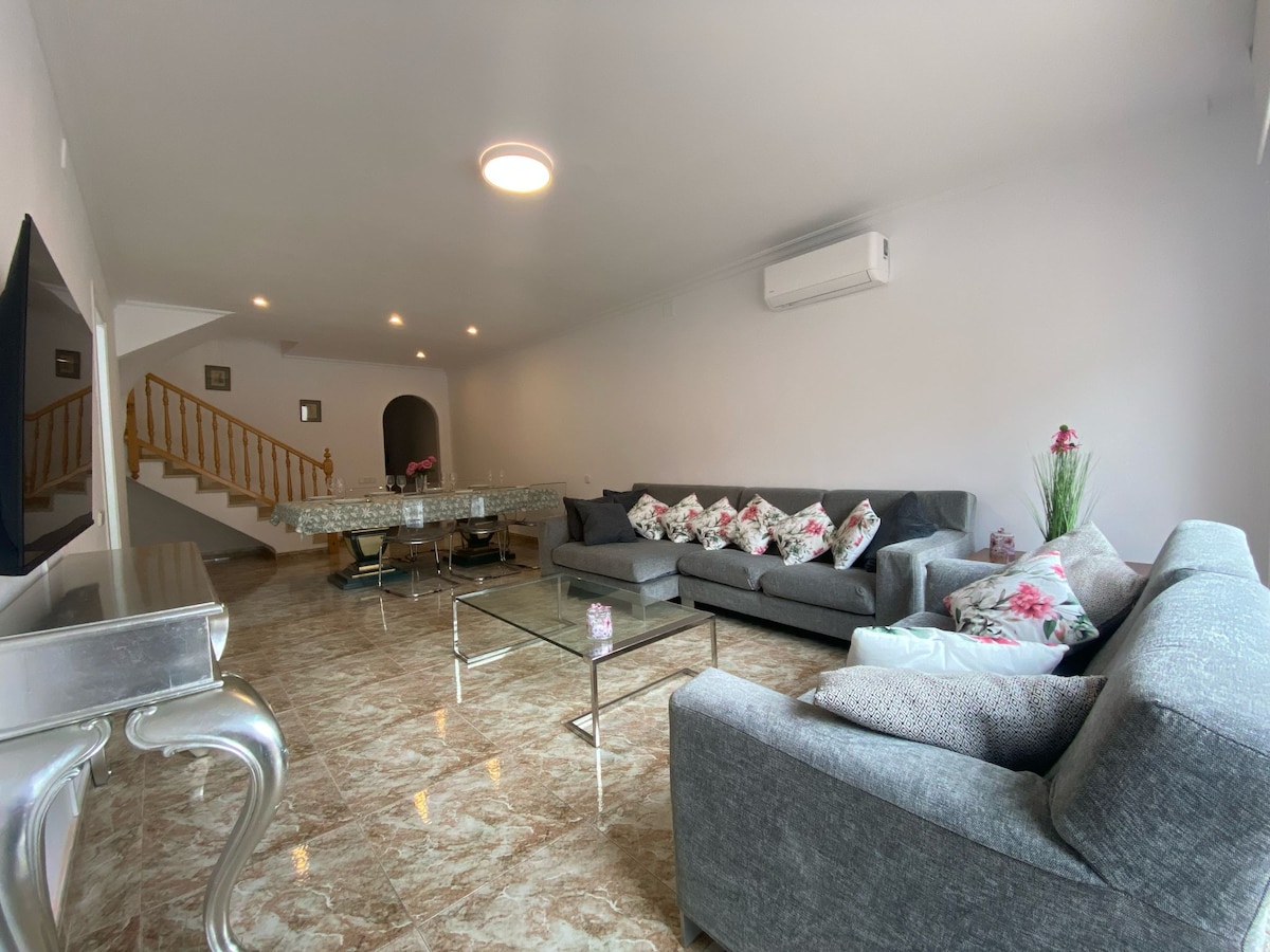 A spacious living area features a gray sofa set adorned with floral accent pillows. A sleek glass dining table sits in the background, complemented by a chandelier overhead. Light-colored walls and tiled flooring enhance the bright and open ambiance. An air conditioning unit is visible on the wall.