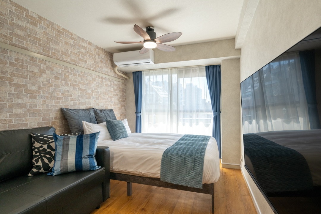 A cozy room featuring a large bed dressed in light linens, accompanied by a dark sofa adorned with decorative cushions. A modern ceiling fan is installed above, and large windows with sheer curtains allow natural light to illuminate the space.